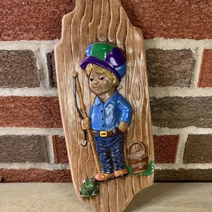 May include: A ceramic wall plaque depicting a boy wearing a purple, green, and blue hat, holding a fishing rod and a bucket. The boy is standing on a wooden plank with a green frog at his feet.