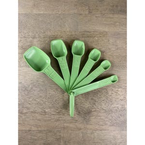 Può includere: Un set di sei cucchiai dosatori verde chiaro a ventaglio su una superficie di legno. Ogni cucchiaio ha una misura diversa, tra cui 0,6 ml, 1,2 ml, 2,5 ml, 5 ml, 15 ml e 59 ml. I cucchiai sono collegati da un anello.