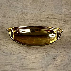 May include: A shiny gold-colored drawer pull with a curved, cup-like design. The hardware has a smooth, reflective surface and is set against a light-colored wooden background. The pull is designed for easy gripping and opening of drawers.