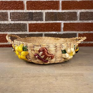 Könnte beinhalten: Gewebter Korb mit Griffen, verziert mit gestickten gelben Blumen und einem braunen Schmetterling. Der Korb besteht aus Naturfasern und hat ein rustikales, handgefertigtes Aussehen. Der Korb hat einen Durchmesser von ca. 25 cm.