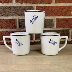 Puede incluir: Tres tazas de cerámica blanca con ribete azul y un diseño de cubiertos azules. Las tazas tienen un asa en el lateral.