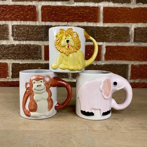May include: Three ceramic mugs with animal designs. One mug features a yellow lion, another a brown monkey, and the third a pink elephant. Each mug has a matching handle and is set against a brick wall.