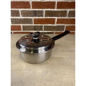 Könnte beinhalten: Edelstahl-Kochtopf mit schwarzem Griff und Deckel mit Backsteinmuster. Der Deckel hat einen schwarzen Knopf. Der Topf steht auf einer hellen Oberfläche, mit einer Backsteinmauer im Hintergrund.