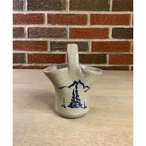 May include: A small, speckled ceramic basket with a handle. The basket features a blue mountain and tree design. The basket is sitting on a light-colored wooden surface, with a brick wall in the background.