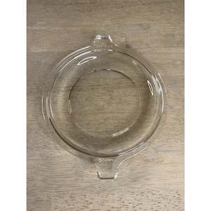 May include: A clear, round glass lid with two small handles. The lid is designed to fit a round dish. The glass is transparent, allowing visibility of the dish's contents. The lid is likely for a casserole dish or similar cookware.