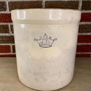 May include: Off-white stoneware crock with a blue crown emblem and the number 3. The antique-style container has a wide mouth and a rounded body. The crock is likely used for storage or decoration.