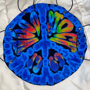 May include: A blue and black tie-dye peace sign with the text "1969" in the centre. The peace sign has a tie-dye pattern in shades of blue, yellow, red, and orange. The peace sign is attached to a black cord.