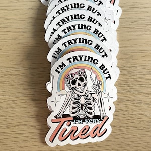 May include: A stack of white round stickers with a skeleton wearing sunglasses and a rainbow behind it. The text on the sticker says "I'm trying but I'm very tired".