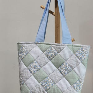May include: A quilted tote bag featuring a diamond pattern of alternating fabric squares in sage green, white with thin stripes, and a floral print with small blue flowers on a white background, finished with two light blue fabric shoulder straps.