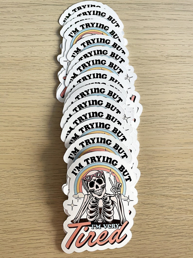 May include: A stack of white round stickers with a skeleton wearing sunglasses and a rainbow behind it. The text on the sticker says "I'm trying but I'm very tired".
