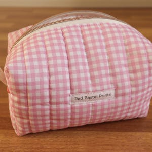 May include: A quilted cosmetic bag featuring a pink and white gingham check pattern. The bag has a top zip closure and a small white fabric label on the front with the text Red Pastel Prints printed in black. The structure is soft and padded.