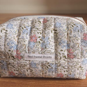 May include: A quilted fabric pouch featuring a floral pattern with light blue, pink, and green botanical elements on a white background. The rectangular bag has a top zip closure and a white rectangular label with the text Red Pastel Prints.