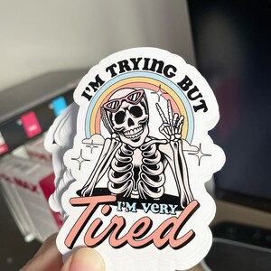May include: A white sticker with a skeleton wearing sunglasses and a rainbow behind it. The text on the sticker reads "I'm trying but I'm very tired".