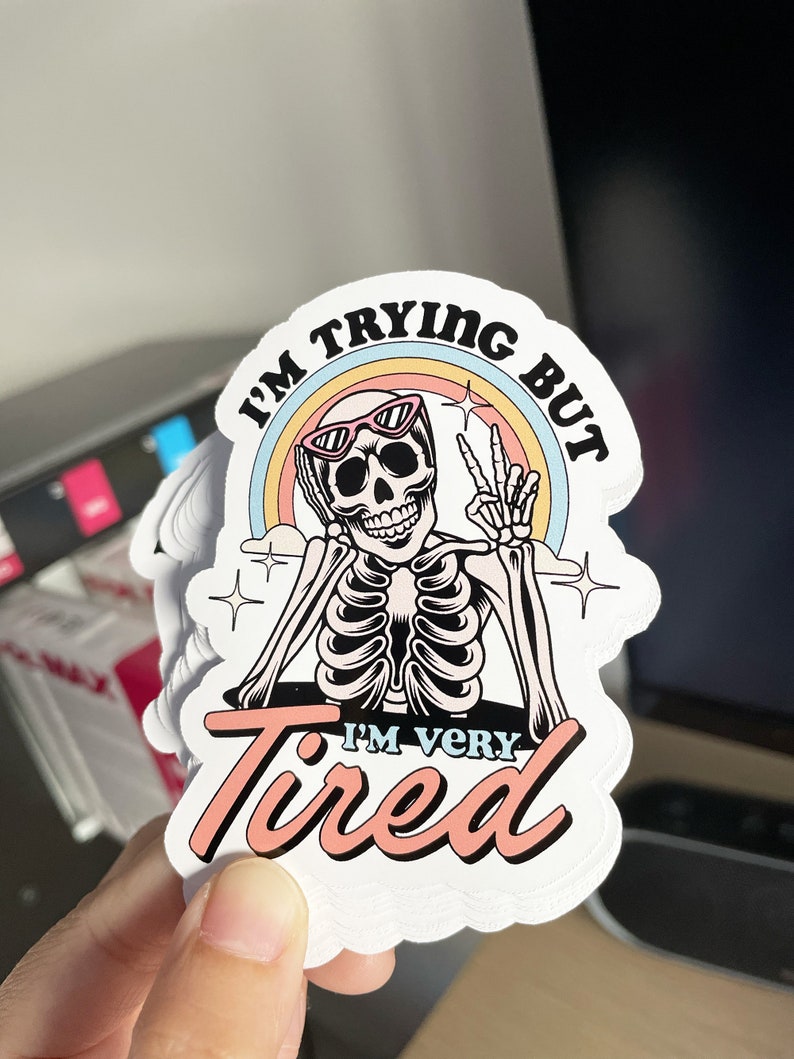 May include: A white sticker with a skeleton wearing sunglasses and a rainbow behind it. The text on the sticker reads "I'm trying but I'm very tired".