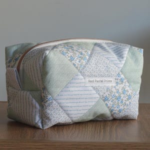 May include: A quilted zipped pouch featuring a patchwork design of light green and white fabrics with small blue floral and striped patterns. A white rectangular fabric label with the text Red Pastel Prints is sewn onto the front surface.