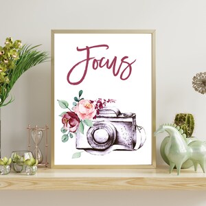 May include: A framed art print featuring a watercolor illustration of a vintage camera with floral accents. The word "Focus" is written in a cursive font above the camera. The print is in a gold frame and is displayed on a wooden shelf.