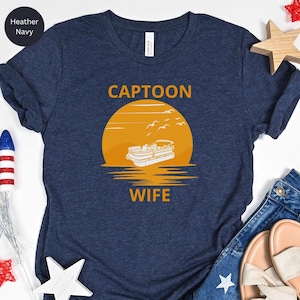 May include: A navy blue t-shirt with the text "CAPTOON WIFE" in white. The design features a yellow sunset with a pontoon boat and birds flying above.
