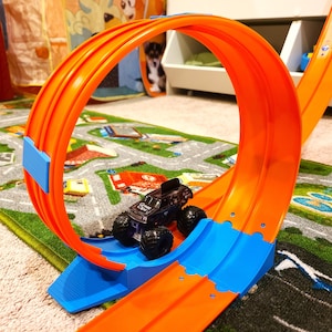 Puede incluir: Pista de carreras de juguete de plástico naranja y azul con un camión monstruo de juguete negro conduciendo por ella.