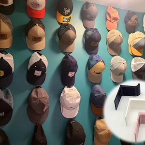 Könnte beinhalten: Eine Wand mit einer Vielzahl von Hüten, die an Haken hängen. Die Hüte sind in verschiedenen Farben und Stilen, darunter Baseballkappen, Bucket-Hats und Beanies. Die Haken sind aus Metall und mit Schrauben an der Wand befestigt. Die Haken sind in Schwarz, Weiß und Rosa erhältlich.