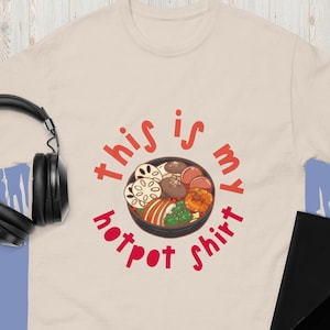 May include: A cream-colored t-shirt featuring a cartoon illustration of a hot pot dish in a black bowl. The text "this is my hotpot shirt" is in red, encircling the bowl. The shirt is made of a soft material.