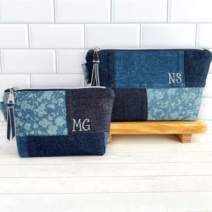 May include: Two zippered pouches made from patchwork denim fabric in varying shades of blue. The smaller pouch features the embroidered letters MG, while the larger pouch displays the letters NS. Both items have silver zippers and denim pull tabs.