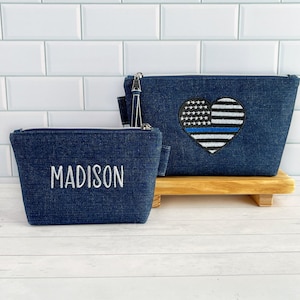 May include: Two denim makeup bags. One bag is personalized with the name "MADISON" in white embroidery. The other bag features a blue line American flag heart in white embroidery.