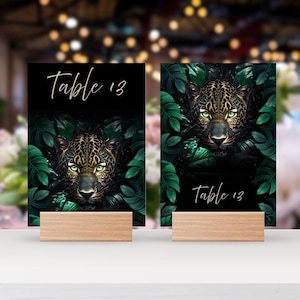 May include: Two table number cards with a jaguar design. The cards feature a black background with a jaguar surrounded by green leaves. The text "Table 13" is written in a cursive font. The cards are held by wooden stands.