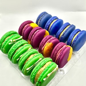 May include: A clear plastic container filled with colorful macarons. The macarons are arranged in rows, with green, purple, and blue macarons visible. The macarons are decorated with white frosting and gold flakes.