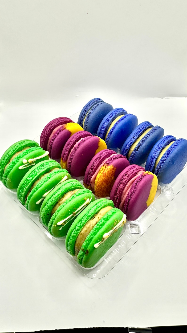 May include: A clear plastic container filled with colorful macarons. The macarons are arranged in rows, with green, purple, and blue macarons visible. The macarons are decorated with white frosting and gold flakes.