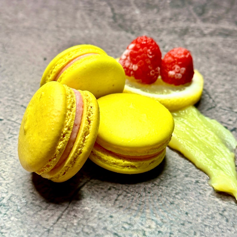 May include: Three yellow macarons with pink filling, topped with two red raspberries and a lemon slice. The macarons are on a gray background.