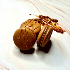 May include: Three brown macarons with caramel drizzle on a white marble surface. The macarons are arranged in a semi-circle with a dollop of caramel sauce behind them.
