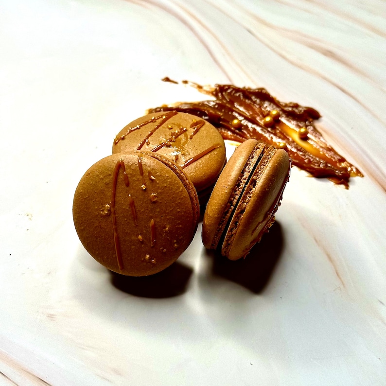 May include: Three brown macarons with caramel drizzle on a white marble surface. The macarons are arranged in a semi-circle with a dollop of caramel sauce behind them.