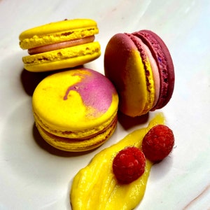 May include: Three yellow macarons with pink and purple swirls, two of the macarons are on a white surface and one is on top of the other two. There is a dollop of yellow lemon curd with two red raspberries on top.