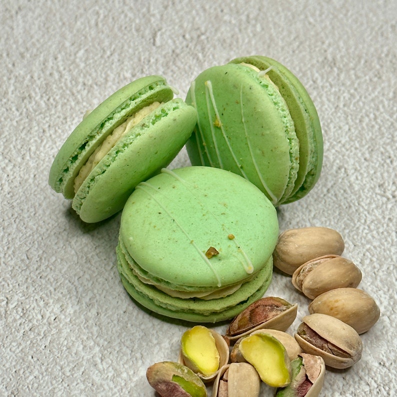 May include: Three green pistachio macarons with white frosting and pistachio nuts scattered around them.