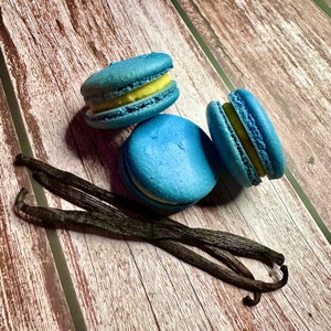 May include: Three blue macarons with yellow filling, arranged on a wooden surface with three vanilla beans.