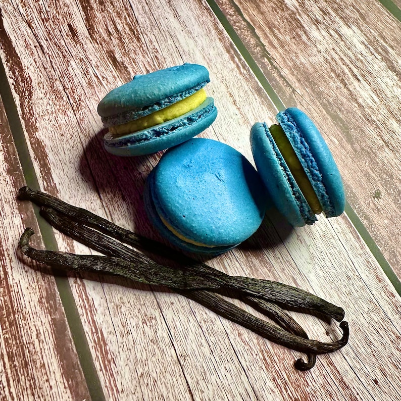 May include: Three blue macarons with yellow filling, arranged on a wooden surface with three vanilla beans.