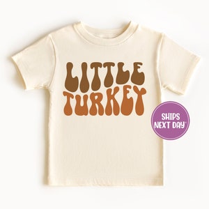 Pu&ograve; includere: Una maglietta color crema con le parole "LITTLE TURKEY" in lettere marroni e arancioni. Le lettere sono in un font retr&ograve; e ondulato.