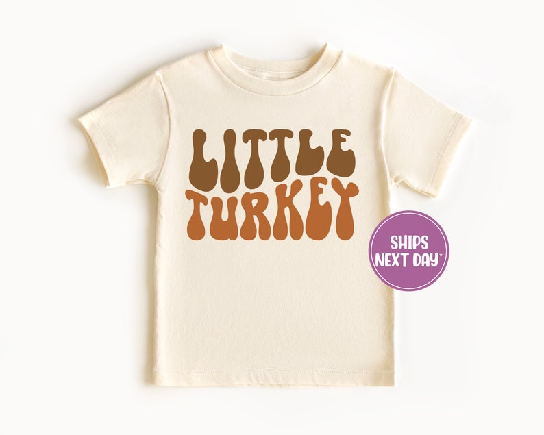 Pu&ograve; includere: Una maglietta color crema con le parole "LITTLE TURKEY" in lettere marroni e arancioni. Le lettere sono in un font retr&ograve; e ondulato.