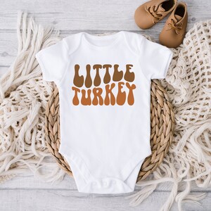 Pu&ograve; includere: Body bianco per neonato con il testo "LITTLE TURKEY" in font retr&ograve; marrone e arancione.