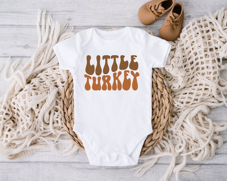 Pu&ograve; includere: Body bianco per neonato con il testo "LITTLE TURKEY" in font retr&ograve; marrone e arancione.