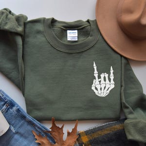 May include: An olive green crewneck sweatshirt featuring a white graphic of a skeleton hand making a rock and roll gesture on the chest. The garment has long sleeves and a ribbed collar, with a Gildan brand label visible at the inner neckline.
