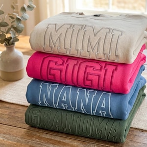 May include: A stack of four crewneck sweatshirts in beige, hot pink, dusty blue, and olive green. Each sweatshirt features large, embroidered block lettering on the front, displaying the words MIMI, GIGI, NANA, and GRANDMA in a matching thread colour.