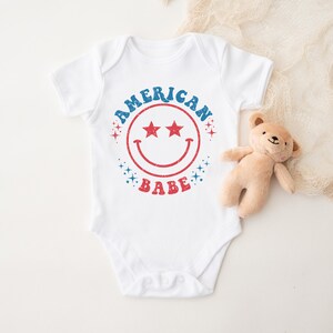 Puede incluir: Un body blanco para beb&eacute; con un gr&aacute;fico rojo, blanco y azul que dice "American Babe" con una cara sonriente y estrellas.