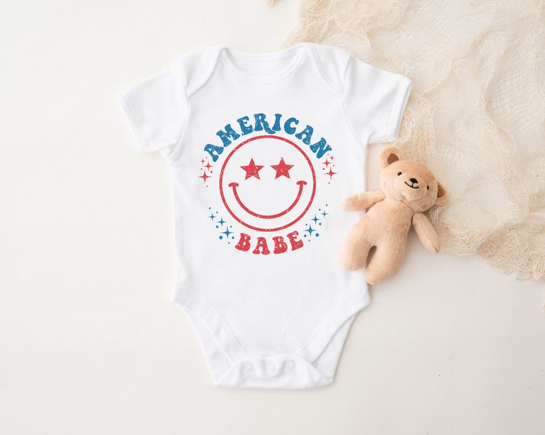 Puede incluir: Un body blanco para beb&eacute; con un gr&aacute;fico rojo, blanco y azul que dice "American Babe" con una cara sonriente y estrellas.