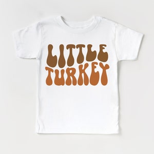 Pu&ograve; includere: Maglietta bianca con il testo "Little Turkey" in marrone e arancione in un font retr&ograve;.