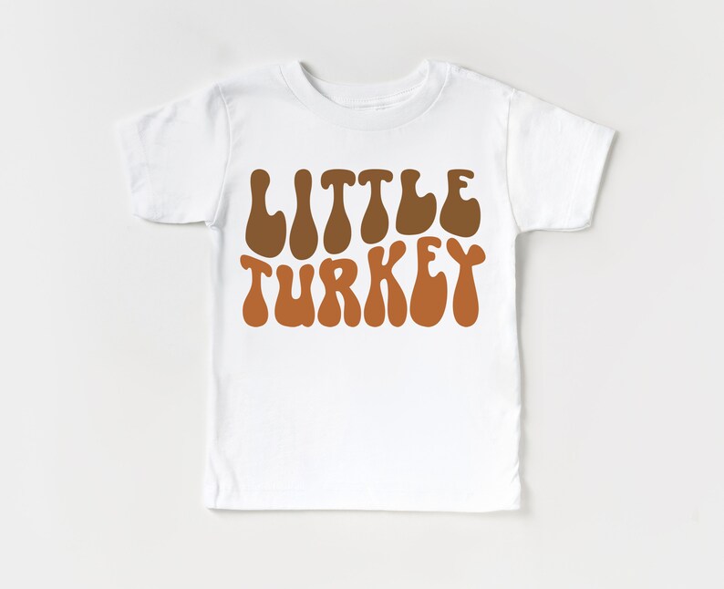 Pu&ograve; includere: Maglietta bianca con il testo "Little Turkey" in marrone e arancione in un font retr&ograve;.