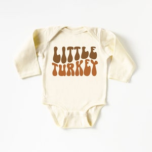 Pu&ograve; includere: Un body per neonato a maniche lunghe di colore crema con il testo "LITTLE TURKEY" in lettere marroni e arancioni in un font retr&ograve;.