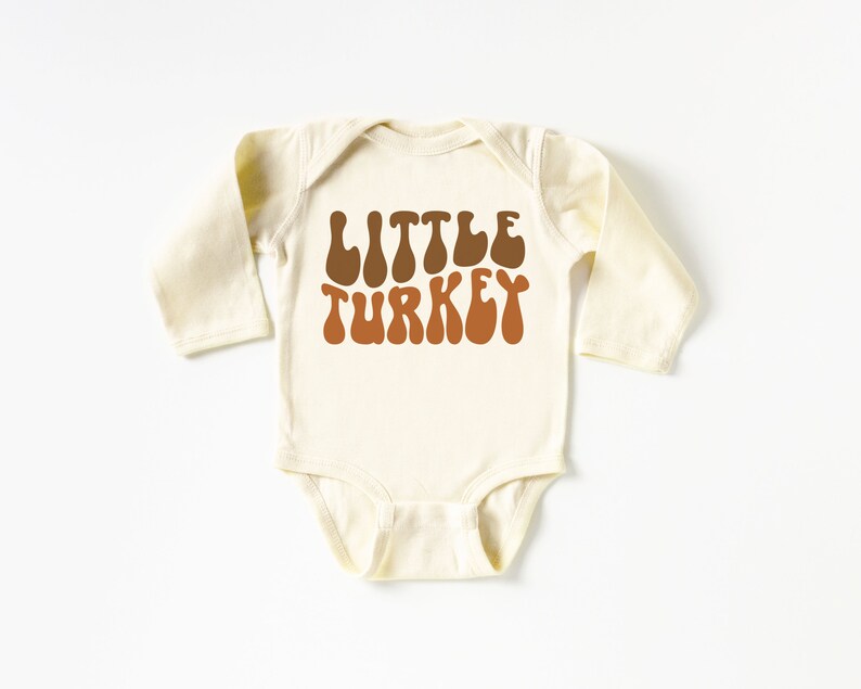 Pu&ograve; includere: Un body per neonato a maniche lunghe di colore crema con il testo "LITTLE TURKEY" in lettere marroni e arancioni in un font retr&ograve;.