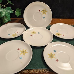 Peut inclure: Ensemble de six soucoupes en céramique blanche. Chaque soucoupe présente une indentation centrale pour une tasse et est ornée d'un motif floral jaune, agrémenté de détails en forme d'étoiles rouges et bleues. Les soucoupes sont disposées sur une surface verte.