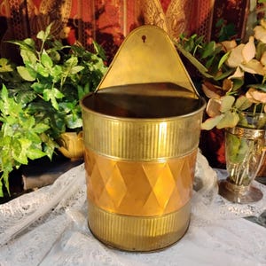Puede incluir: Una maceta de pared de latón y cobre con forma cilíndrica y textura acanalada en las secciones superior e inferior. La banda central muestra un patrón de diamantes en relieve. Incluye una placa trasera puntiaguda con un orificio para colgar.